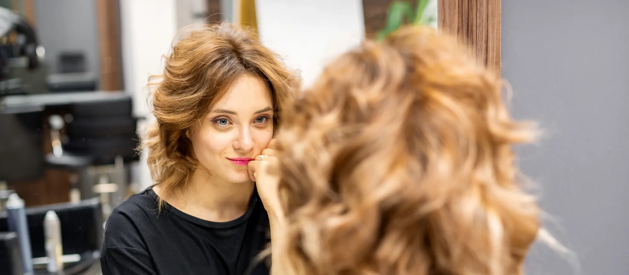 Vrouw met schouderlang, golvend lichtbruin haar bekijkt zichzelf glimlachend in de spiegel van een salon.